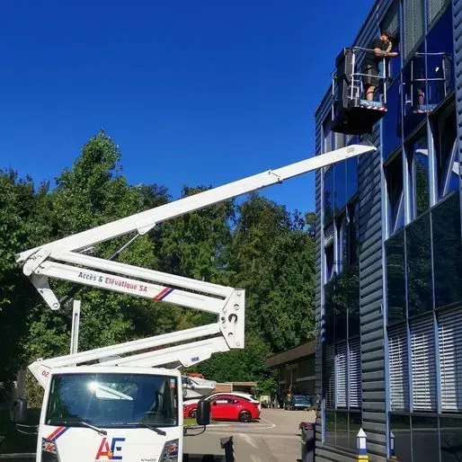 Un ouvrier lave les vitres d'un immeuble moderne dans un ascenseur. Ciel bleu. Chariot élévateur blanc.