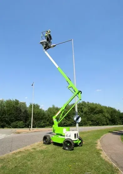 Nacelle élévatrice verte levée, avec une personne dans la nacelle, travaillant sur un lampadaire contre un ciel bleu.