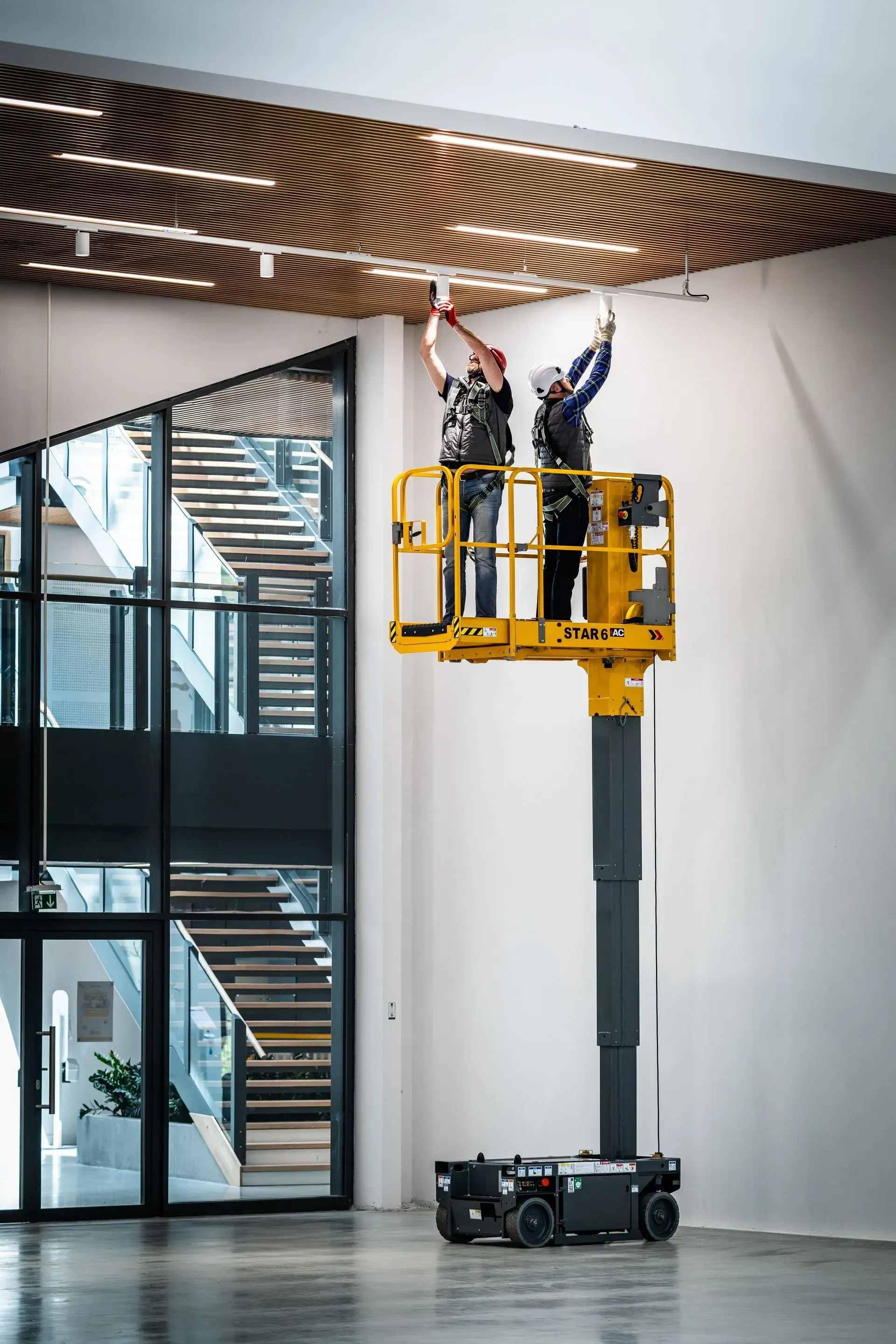 Deux ouvriers sur une nacelle jaune installent des luminaires sur un plafond en bois dans le hall d'un immeuble.