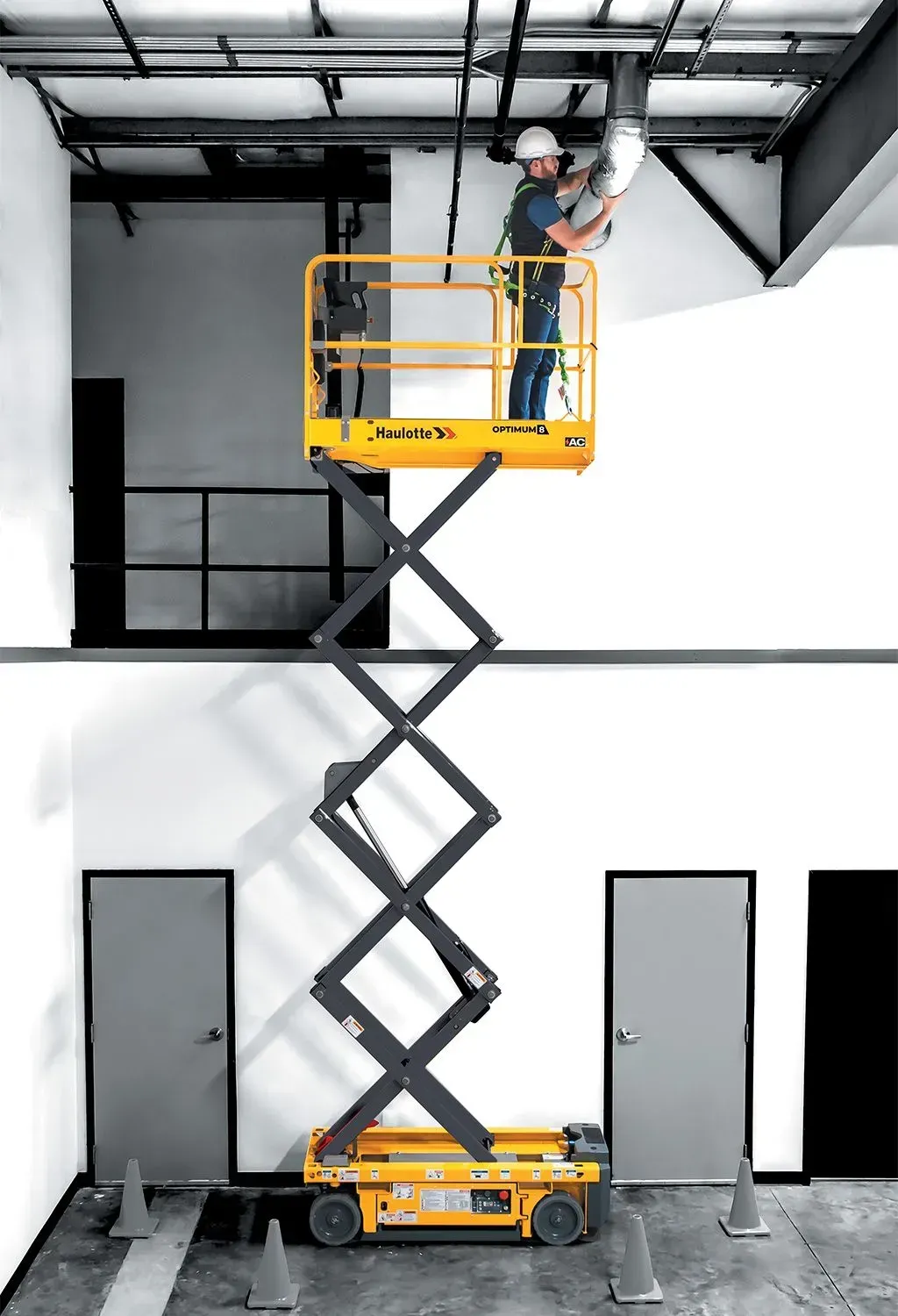 Ouvrier dans une nacelle élévatrice réparant un luminaire suspendu. Ascenseur jaune, portes grises, murs blancs, intérieur.