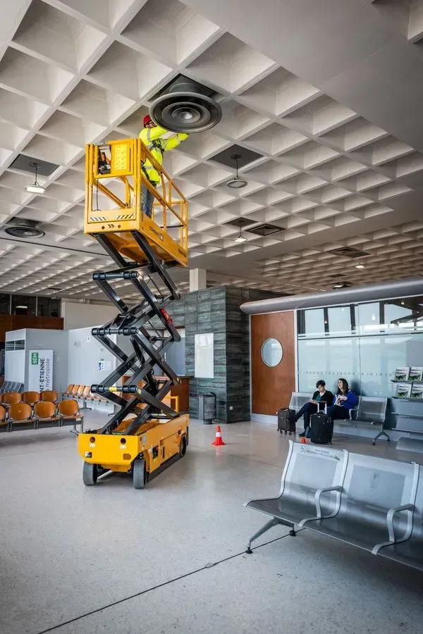 Un employé d'un ascenseur travaille sur une grille d'aération au plafond d'un aéroport. 