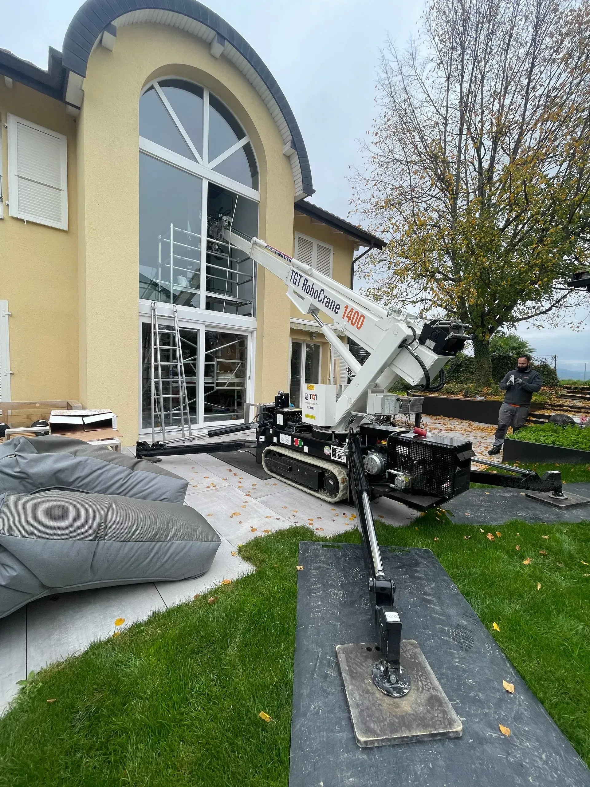 Une grue soulève un grand panneau de verre dans la fenêtre cintrée d'une maison jaune.