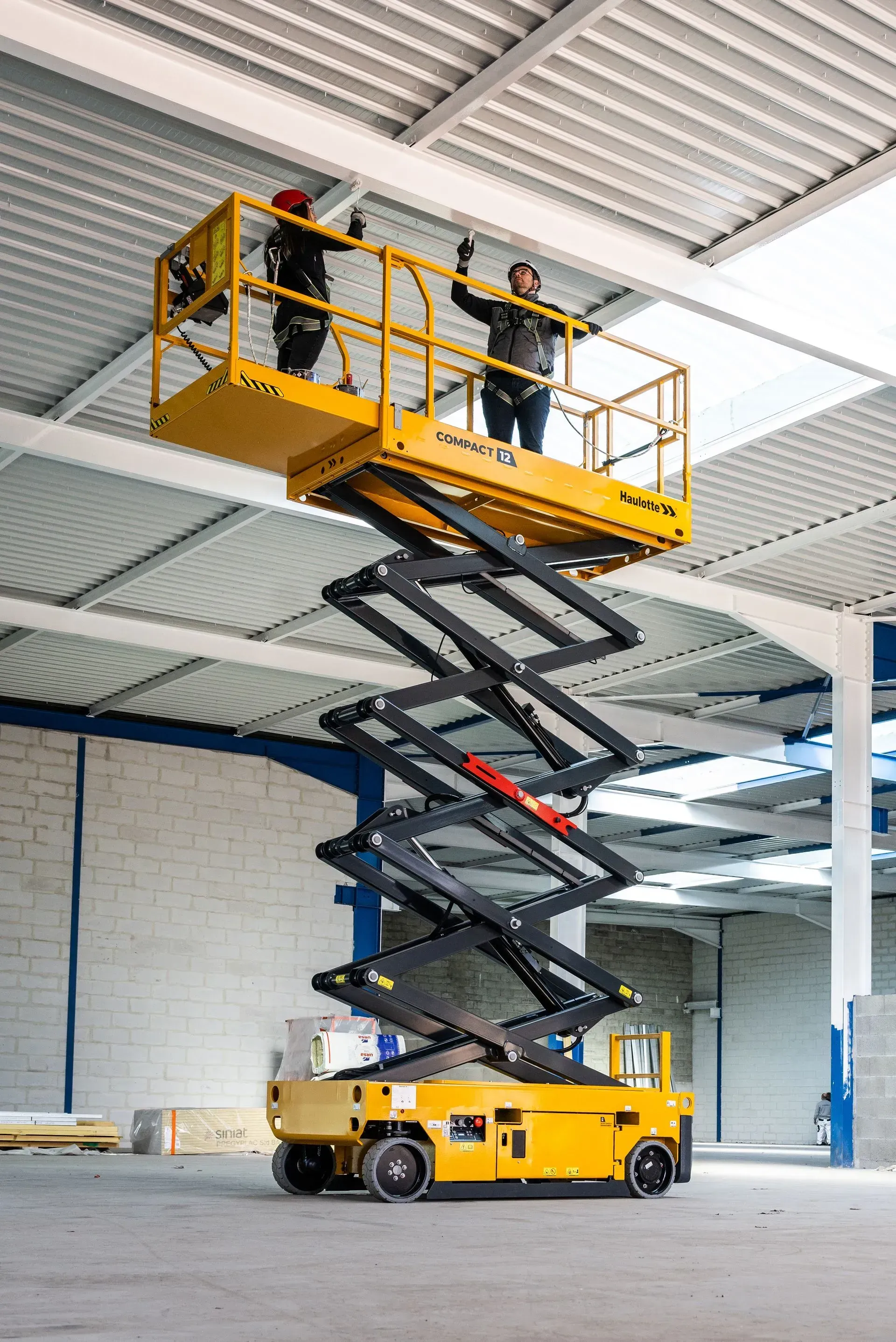 Deux ouvriers sur un élévateur à ciseaux jaune peignent un plafond blanc dans un entrepôt.