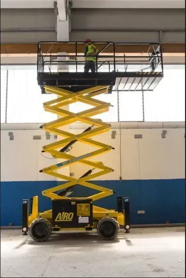 Un élévateur à ciseaux jaune avec un travailleur sur la plate-forme surélevée à l'intérieur d'un bâtiment.