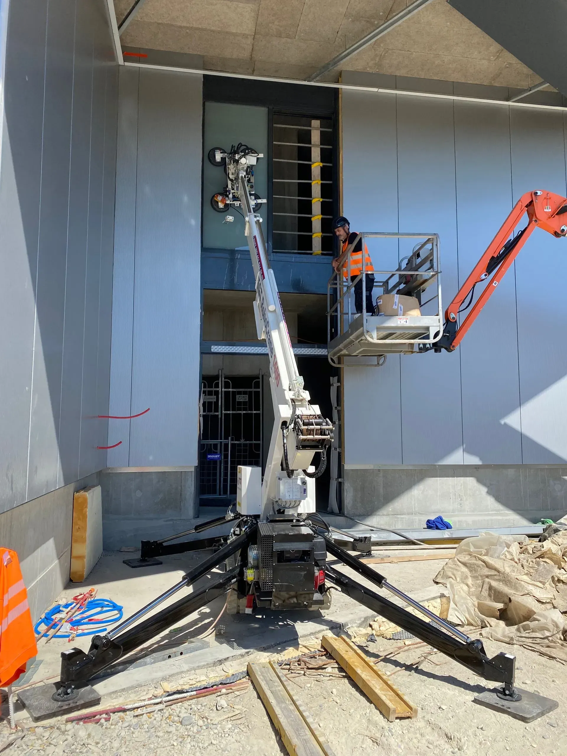 Ouvrier du bâtiment sur un ascenseur installant un revêtement extérieur. Ascenseur blanc, gilet orange, extérieur de jour.