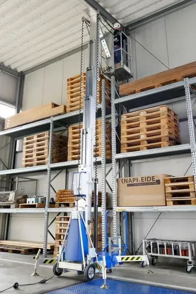 Une personne dans un ascenseur bleu cherchant des objets sur une étagère haute dans un entrepôt.