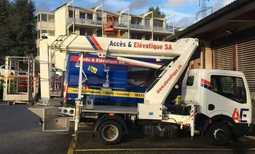 Camion blanc avec grue déployée ; logo « Accès & Élévatique SA ». Bâtiment et parking en arrière-plan.