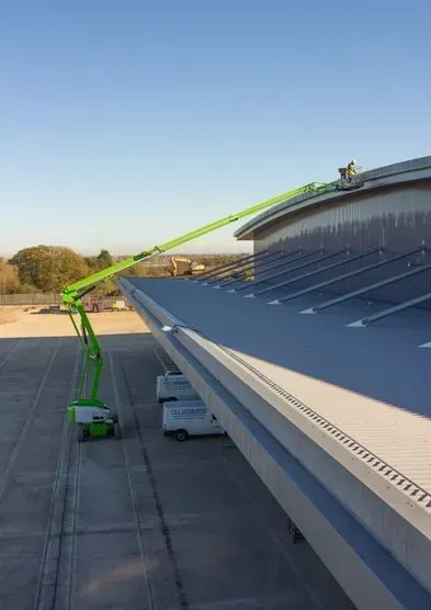 Nacelle élévatrice verte atteignant le toit d'un immeuble. Une personne travaille sur le bord du toit. Ciel dégagé.