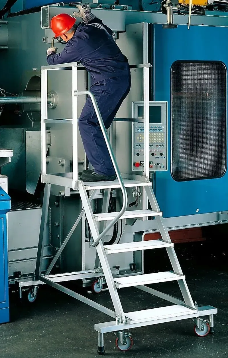 Une personne en salopette monte sur une plate-forme de travail roulante en aluminium à côté de machines dans une usine.