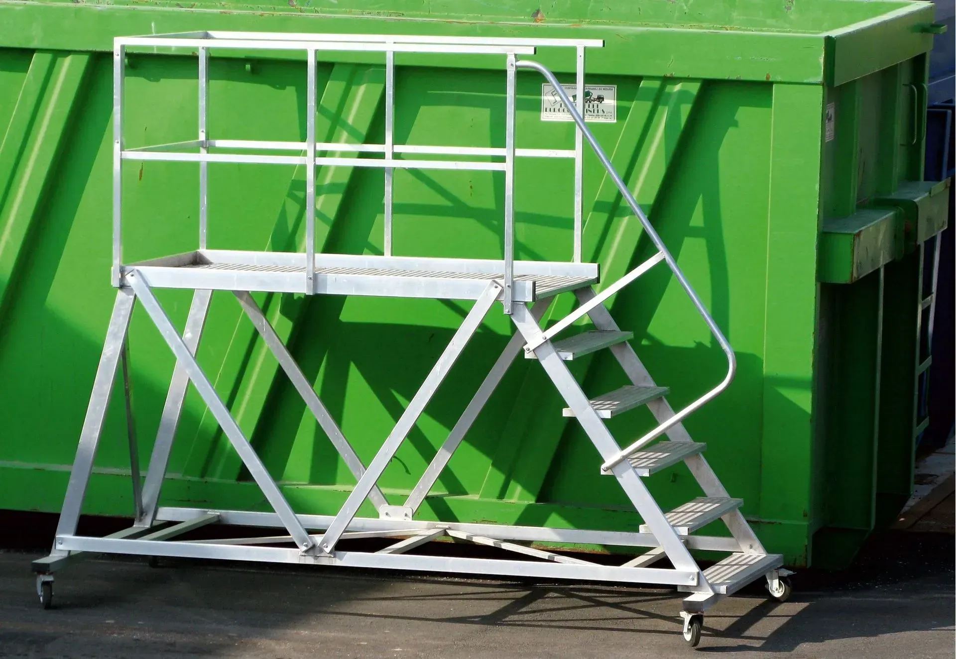 Plateforme de travail en aluminium avec escaliers et garde-corps devant une benne verte.
