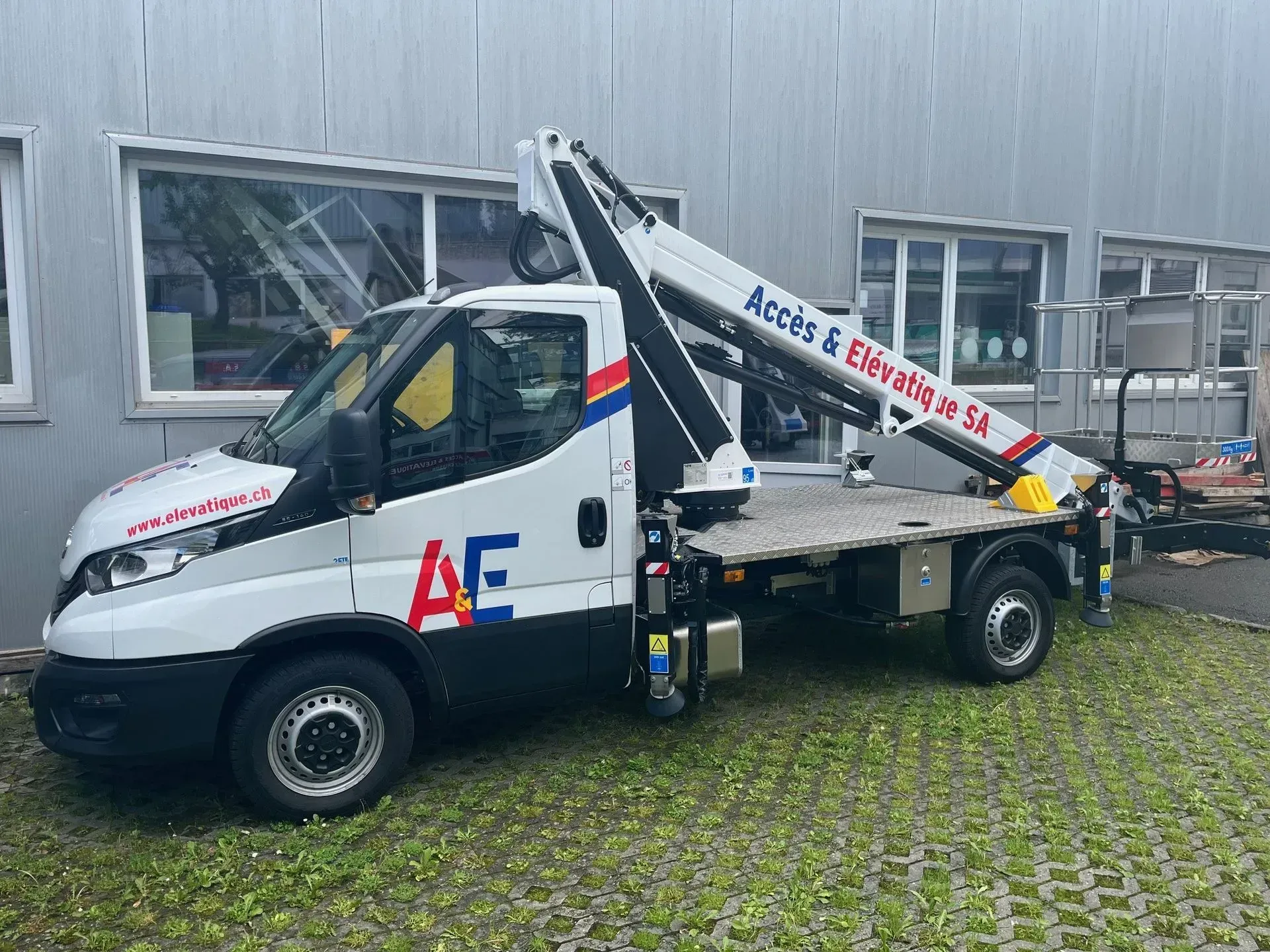 Camion blanc avec nacelle élévatrice; logo « AE » sur le côté; devant le bâtiment.