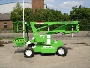 Nacelle élévatrice verte sur roues, prête à être soulevée. En extérieur, près d'un bâtiment et d'arbres.