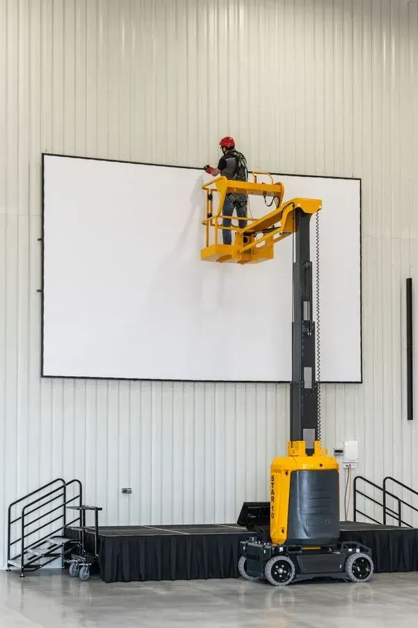 Un ouvrier sur un ascenseur installe un grand écran blanc à l'intérieur. Ascenseur jaune, casque de sécurité rouge.