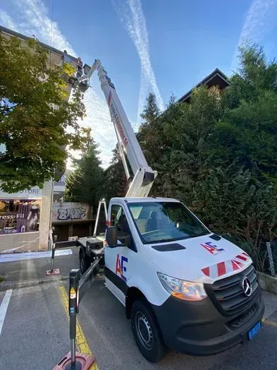 Camion blanc avec nacelle déployée vers un bâtiment, probablement pour des travaux de réparation. Ciel bleu.
