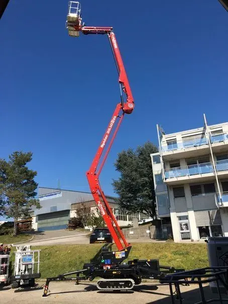 Une nacelle élévatrice articulée rouge, déployée en hauteur, près d'un bâtiment par une journée ensoleillée.