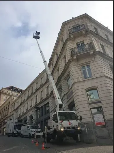 Bras de levage monté sur camion atteignant le coin d'un bâtiment; autres véhicules et cônes dans la rue.