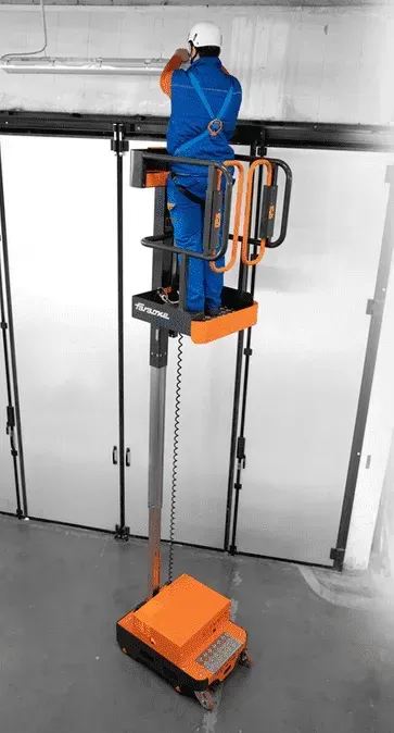 Un homme en salopette bleue sur un ascenseur orange, travaillant sur quelque chose au-dessus d'une porte de garage fermée.