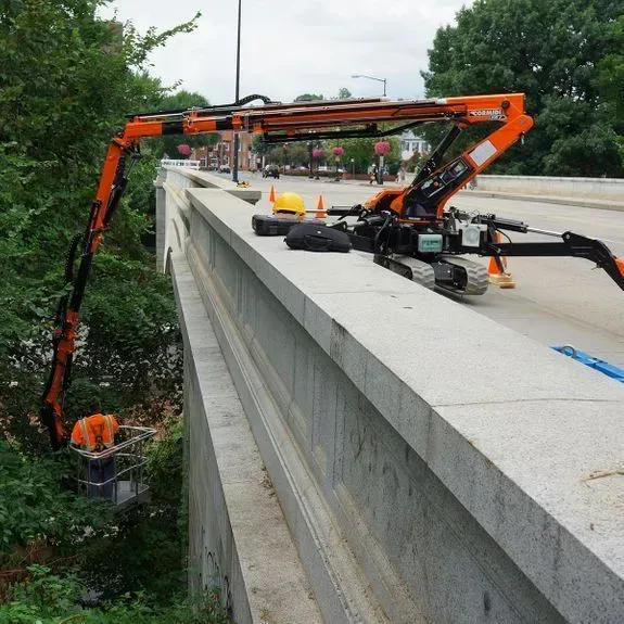 Ascenseur robotisé orange sur un pont de pierre, taillant la verdure ; journée ensoleillée.