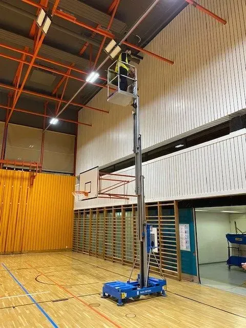Un ouvrier répare les lumières d'un gymnase dans un ascenseur. Le sol, les murs et le panier de basket sont visibles.