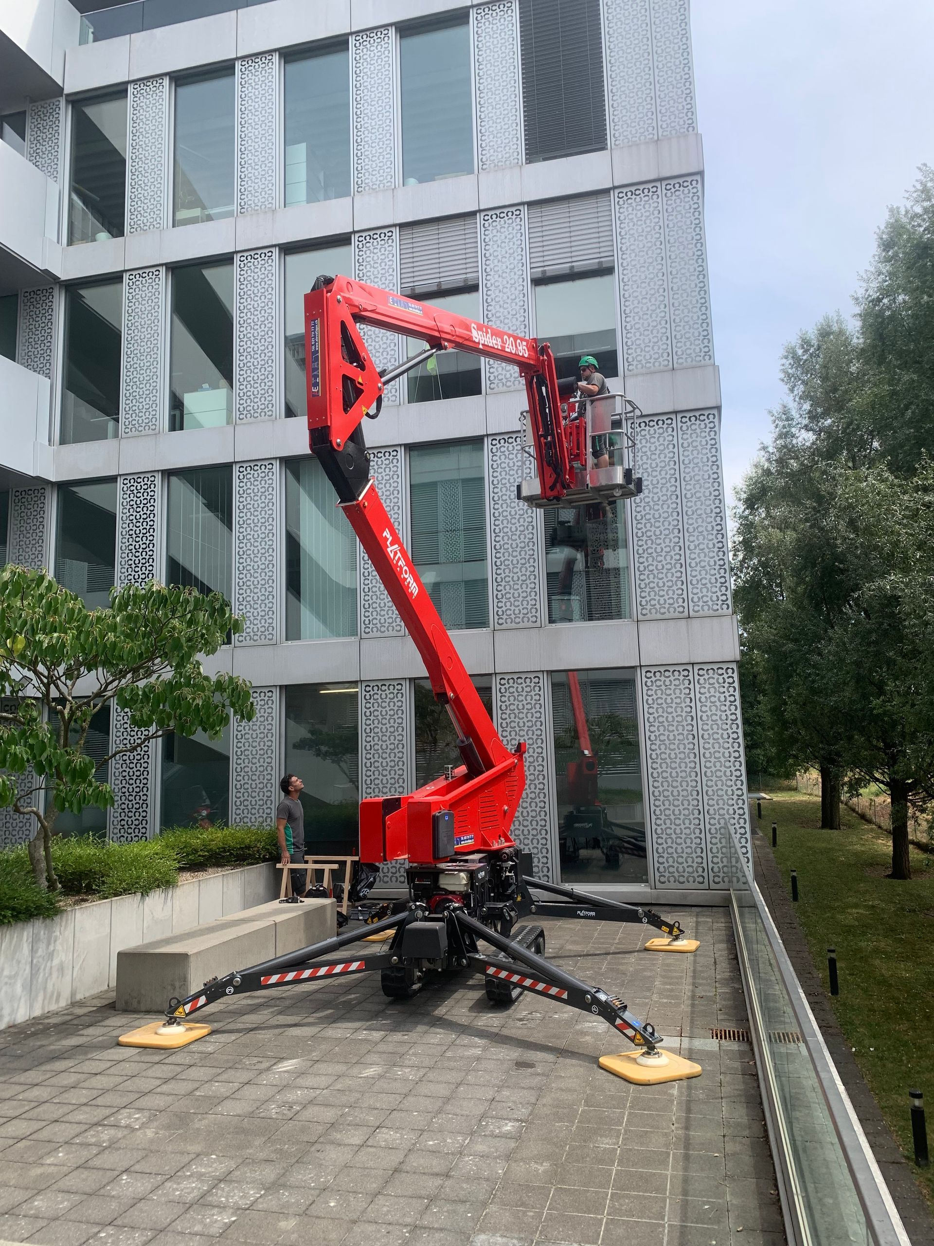 Une nacelle télescopique rouge est garée à côté d'un bâtiment gris dans une cour pavée.