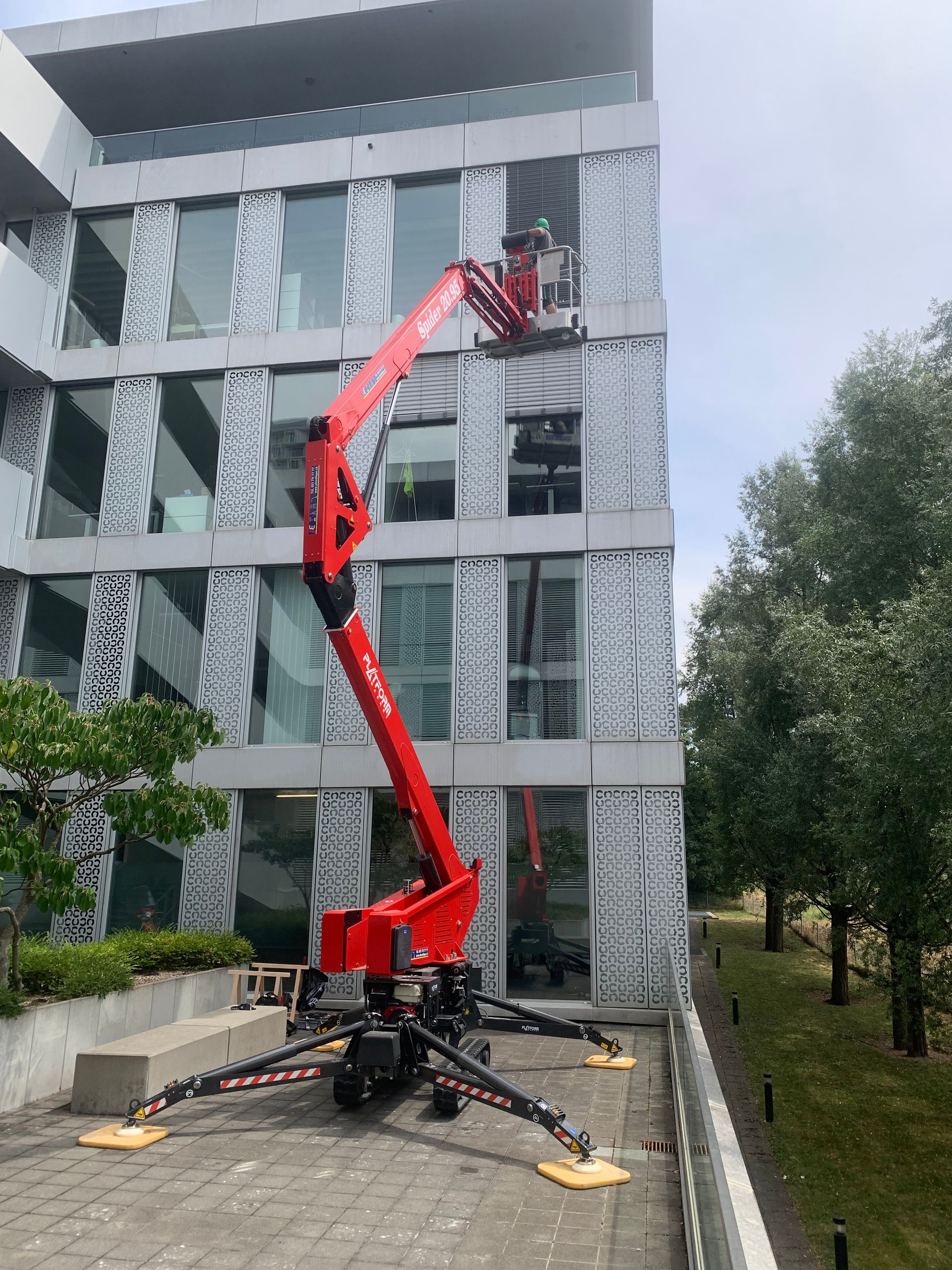 Une nacelle élévatrice articulée rouge, déployée en hauteur, près d'un bâtiment par une journée ensoleillée.