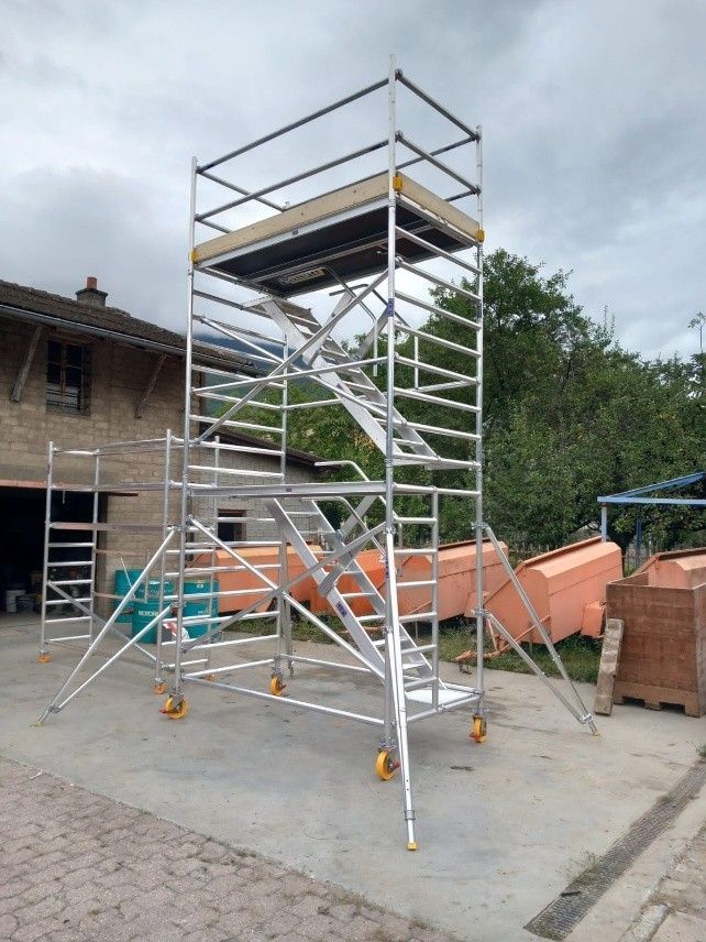 Tour d'échafaudage avec escalier métallique et plateforme sur un chantier de construction, avec des barrières orange à proximité.