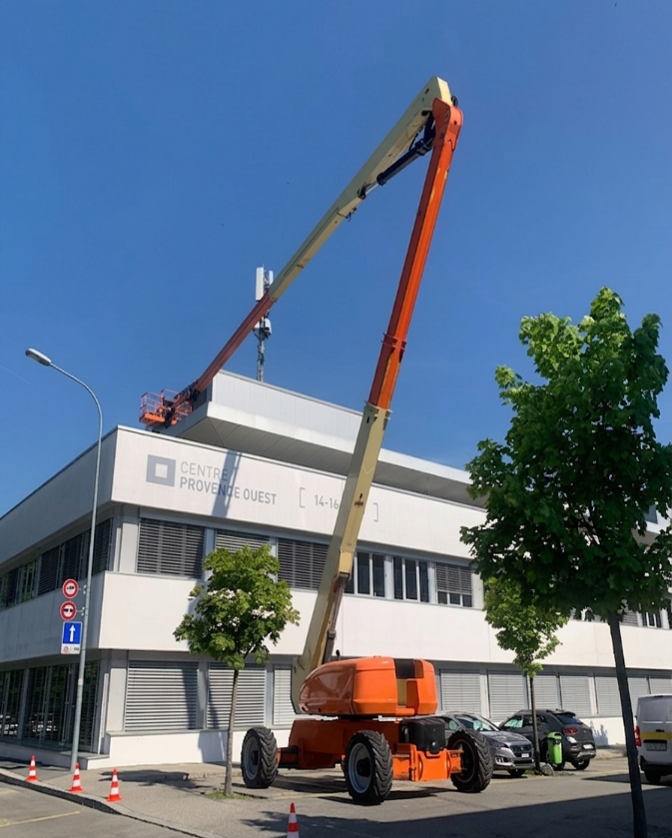 Une nacelle élévatrice orange et beige est déployée à côté d'un bâtiment blanc sous un ciel bleu, des voitures étant garées en contrebas.