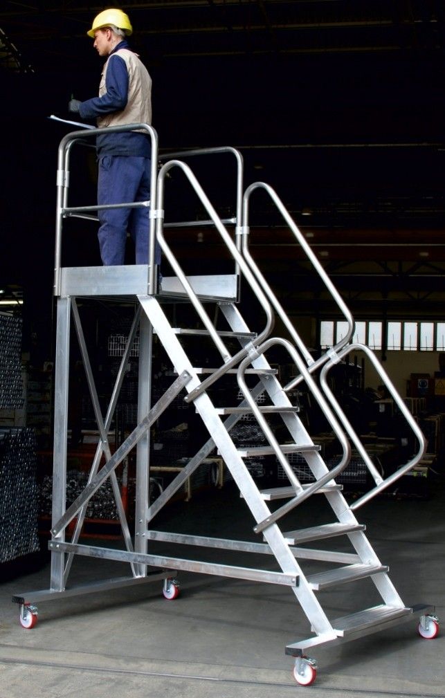 Ouvrier sur une plateforme d'escalier métallique, portant un casque et un équipement de sécurité, dans un entrepôt.