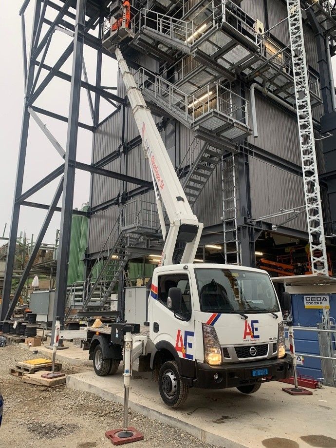 Camion utilitaire blanc avec nacelle élévatrice déployée, stationné à côté d'une structure industrielle et d'un escalier en acier