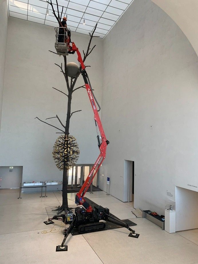 Une grue rouge soulève une grande sculpture d'arbre dans une galerie blanche aux allures de musée, avec un haut plafond voûté.