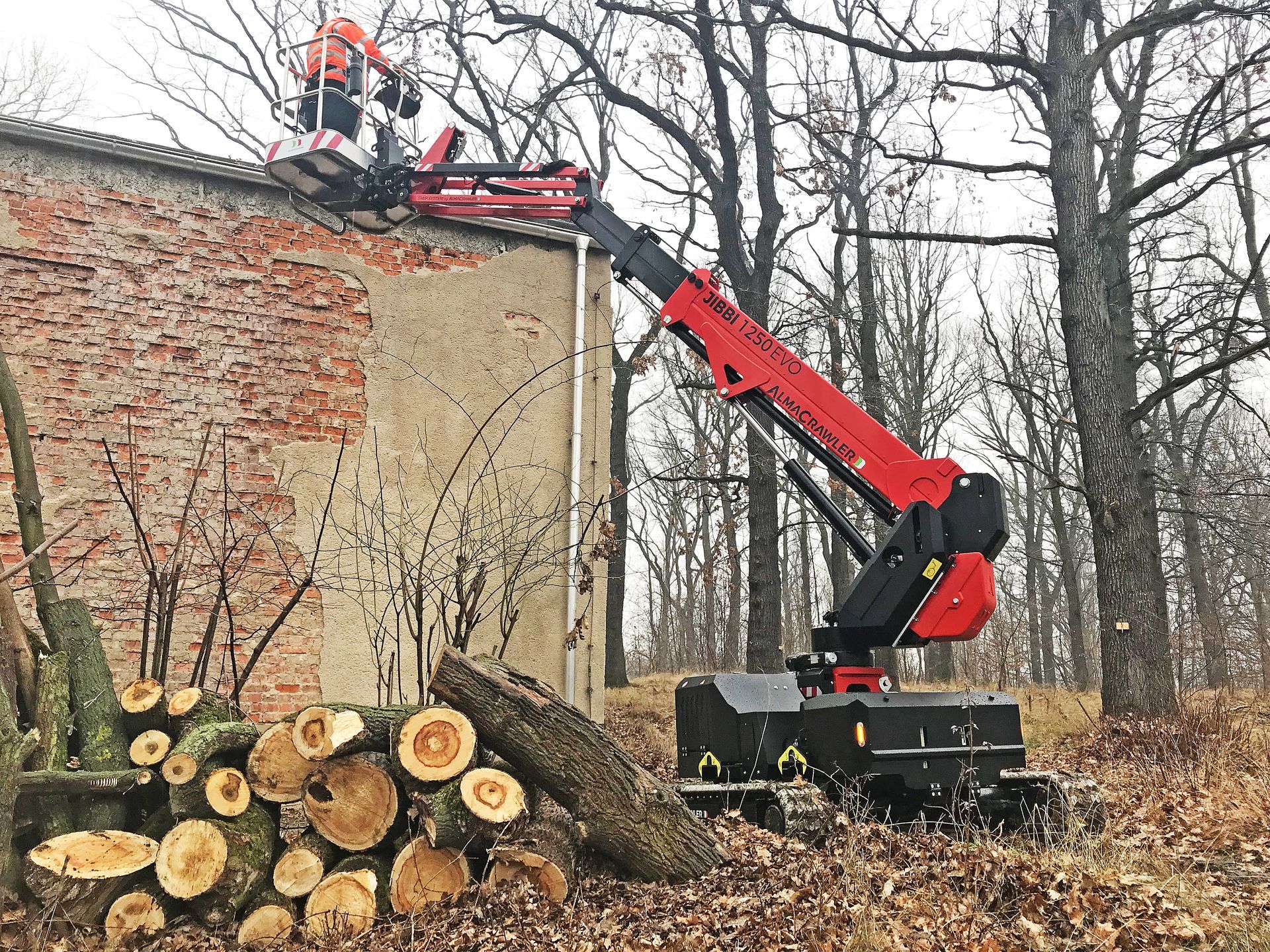 Une grue forestière rouge soulève un tronc d'arbre coupé à côté d'un bâtiment en briques et de grumes empilées dans une zone boisée.