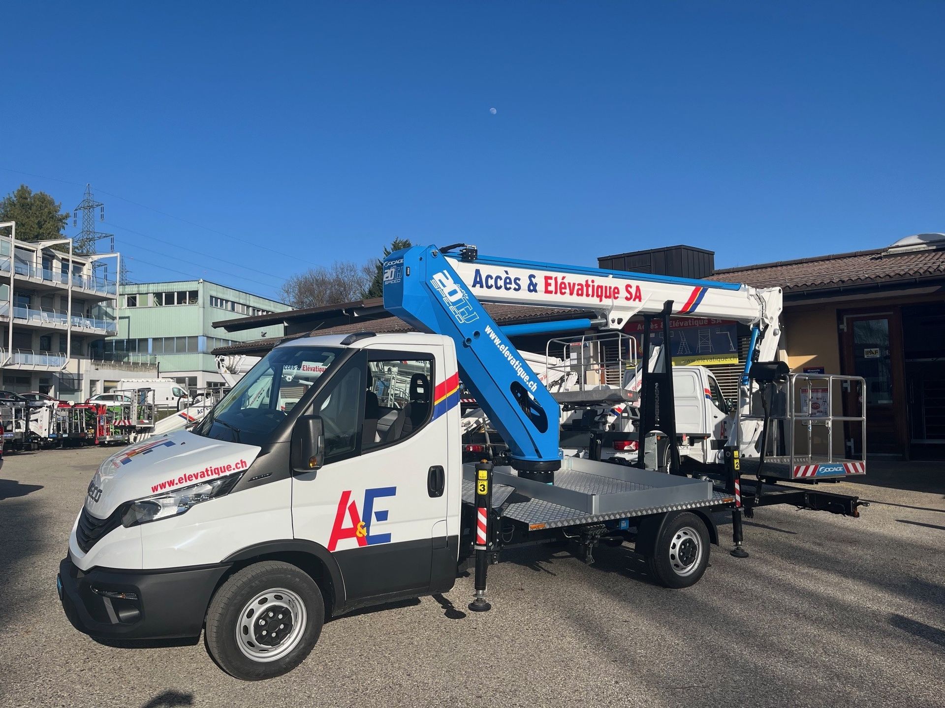 Camion blanc avec nacelle élévatrice; logo « AE » sur le côté; devant le bâtiment.