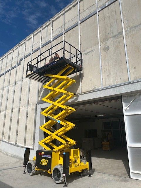 Une nacelle élévatrice jaune est levée contre un mur d'entrepôt, avec un ouvrier dans la nacelle près du toit.