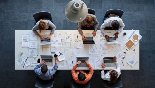 Menschen sitzen um einen Tisch herum und führen unter einer Hängelampe ein Brainstorming mit Laptops und Papier durch.