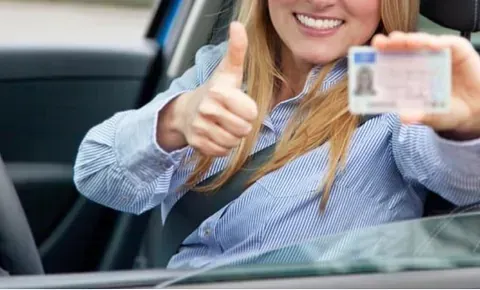 Una mujer está sentada en un automóvil sosteniendo una licencia de conducir y haciendo un gesto de aprobación con el pulgar hacia arriba.