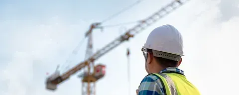 Un hombre que lleva casco y chaleco de seguridad está parado frente a una grúa de construcción.