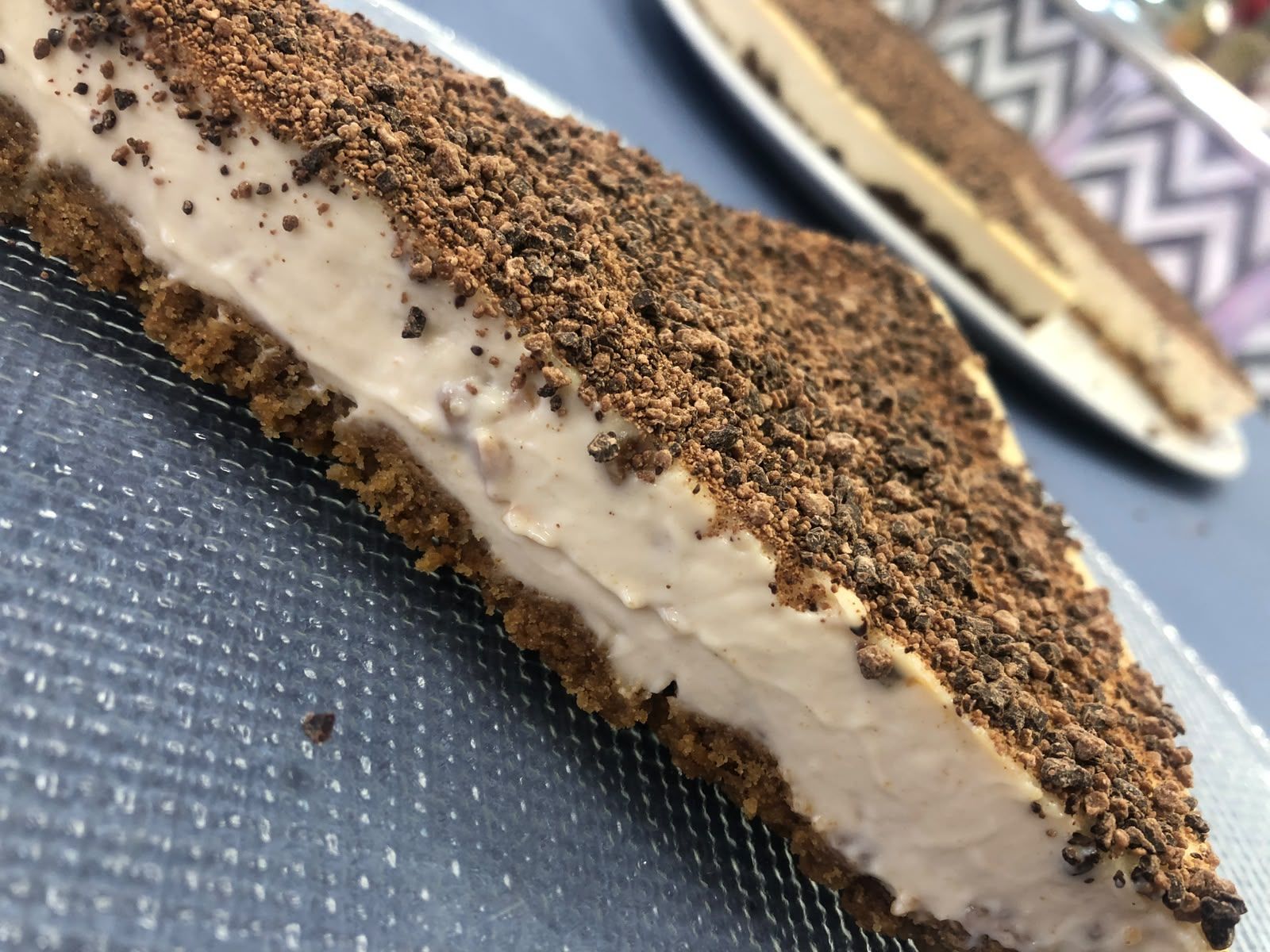 Porción de tarta de queso con cobertura de chocolate en un plato azul, con otra porción desenfocada al fondo