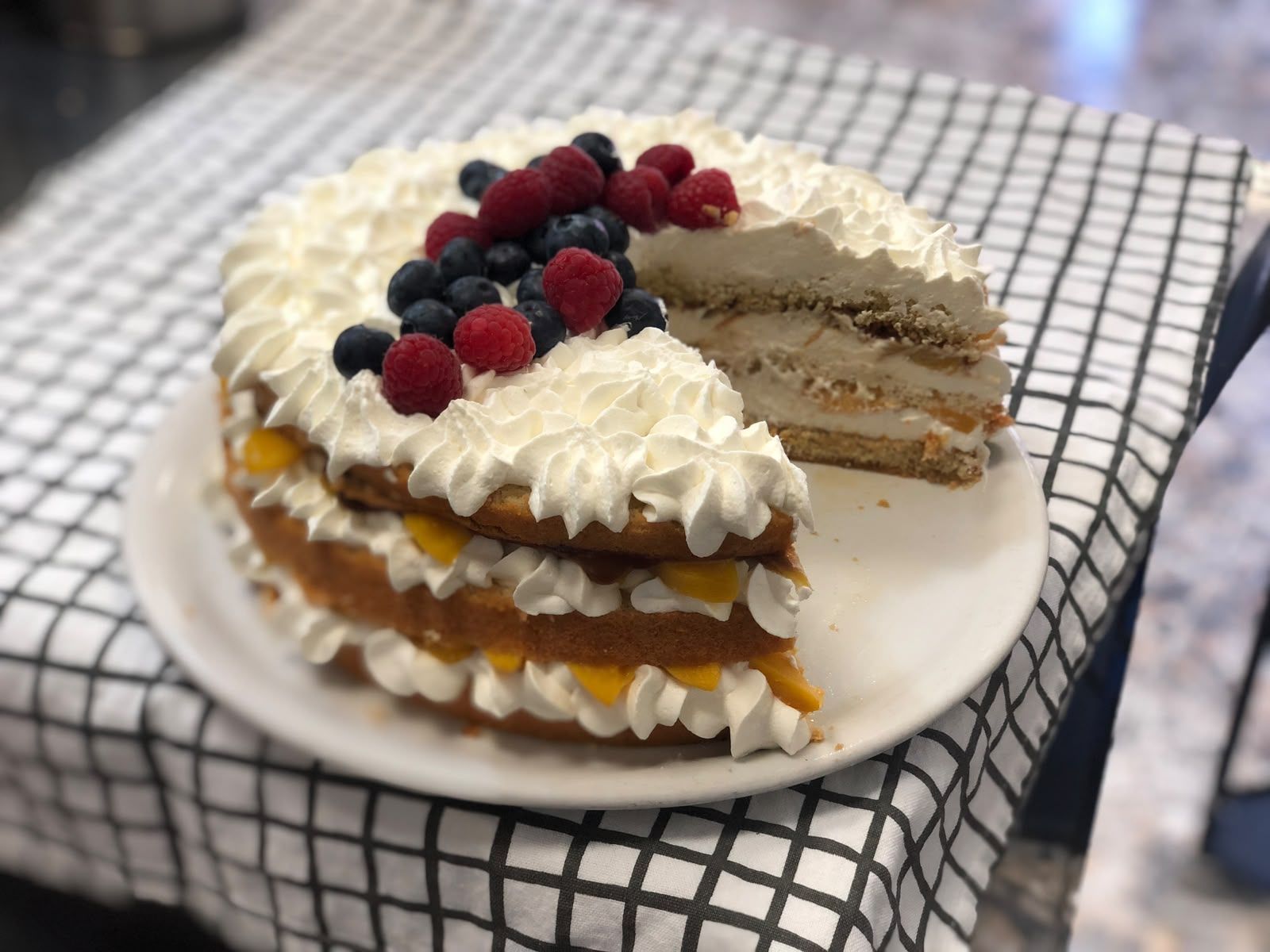 Tarta en capas cubierta de frutos rojos y nata montada sobre un plato blanco, con una porción retirada, sobre un mantel