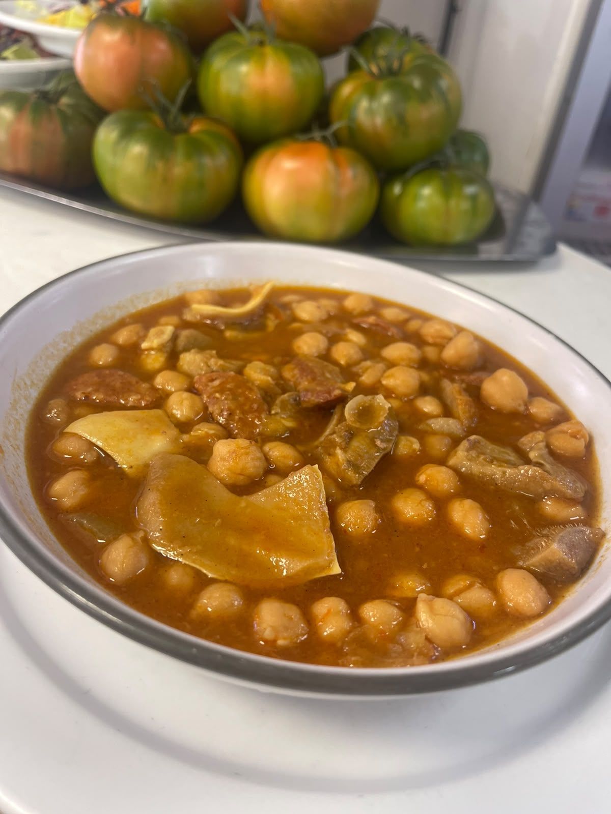 Un plato de guiso de garbanzos con patatas sobre una mesa, con tomates amontonados al fondo.