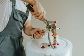 Un plombier en salopette grise serre un tuyau de chauffe-eau à l'aide d'une clé.