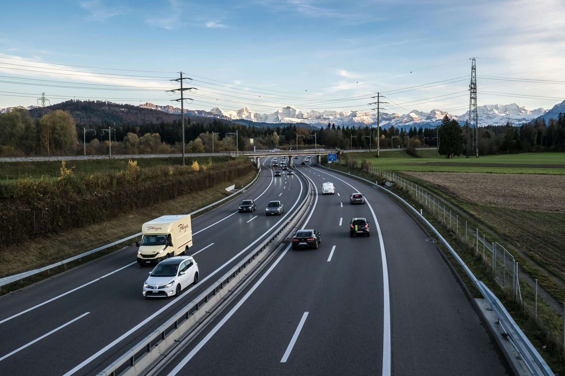 Carretera con coches circulando, cordillera al fondo, cielo azul.