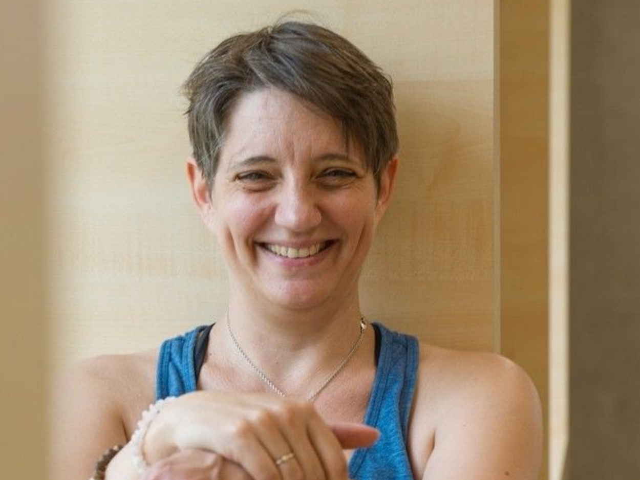 Portraitfoto von Christine Herholz, die lächelt, sie trägt ein blaues Tanktop und lehnt an einer hellen Holzwand.