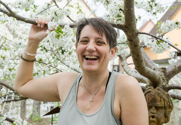 Portrait von Christine Herholz im Sport-Outfit, lachend vor einem blühenden Baum, sie hält einen der Äste in der Hand.