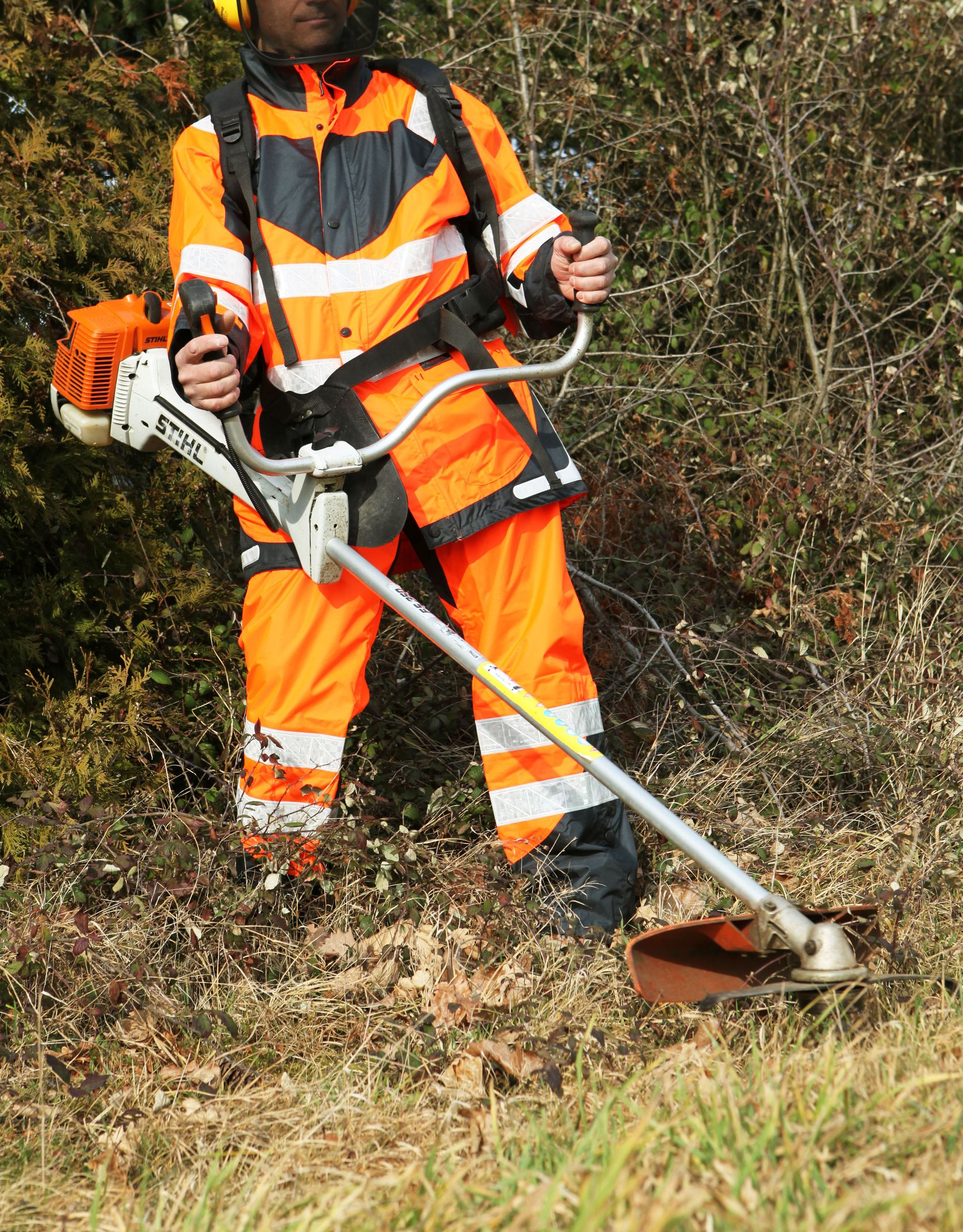 Une personne portant un équipement de sécurité orange utilise une débroussailleuse dans une zone herbeuse.