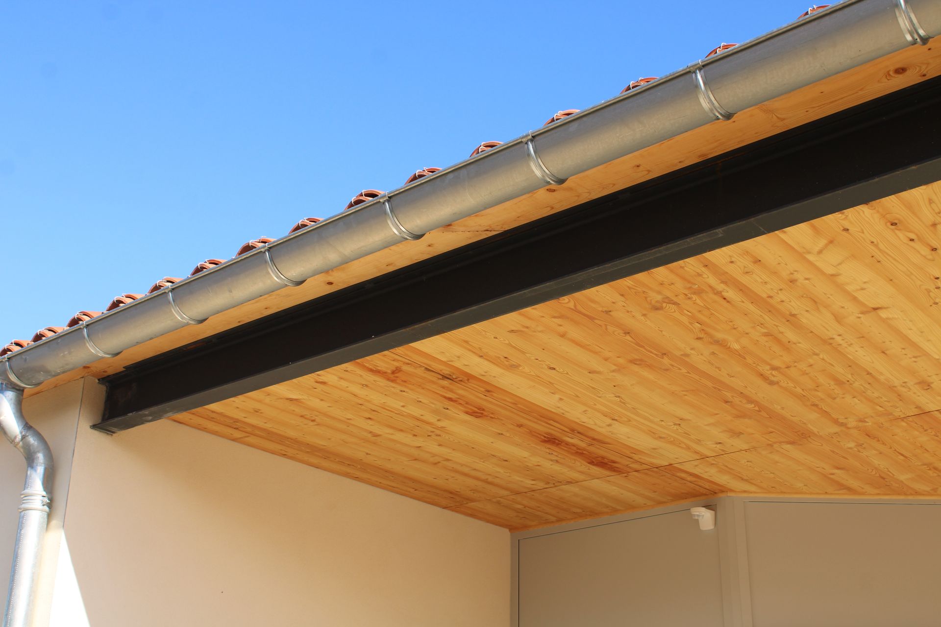 Toit en bois avec une poutre noire et une gouttière grise sur fond de ciel bleu.