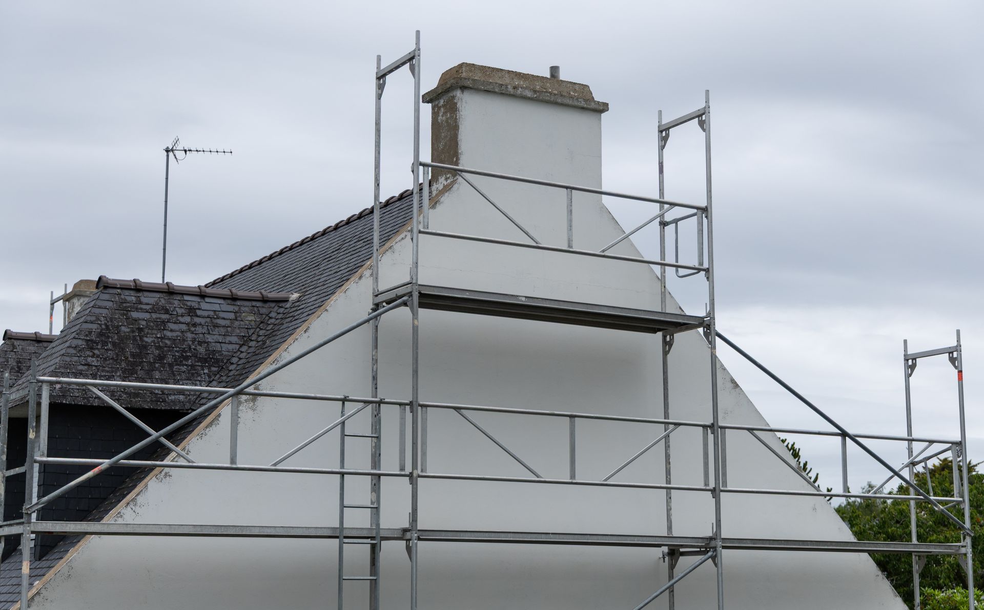 Échafaudage installé devant une maison blanche pour des travaux de cheminée. Toit gris et ciel couvert.