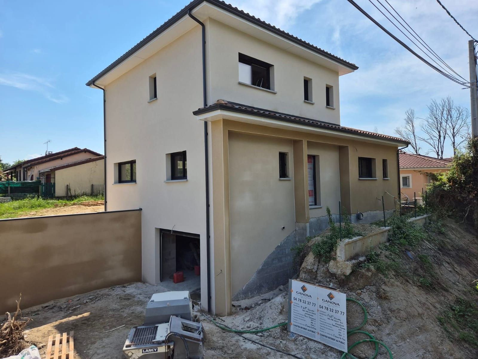 Maison beige de deux étages en construction avec garage, entourée de terre et d'autres maisons par une journée ensoleillée.