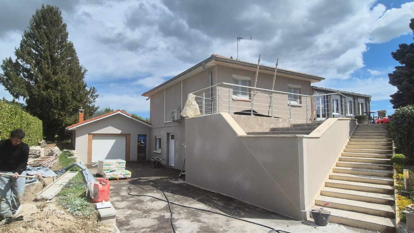 Maison à deux étages avec garage indépendant, rénovation extérieure. Escalier, échafaudage, ciel nuageux. Un homme se trouve près du chantier.