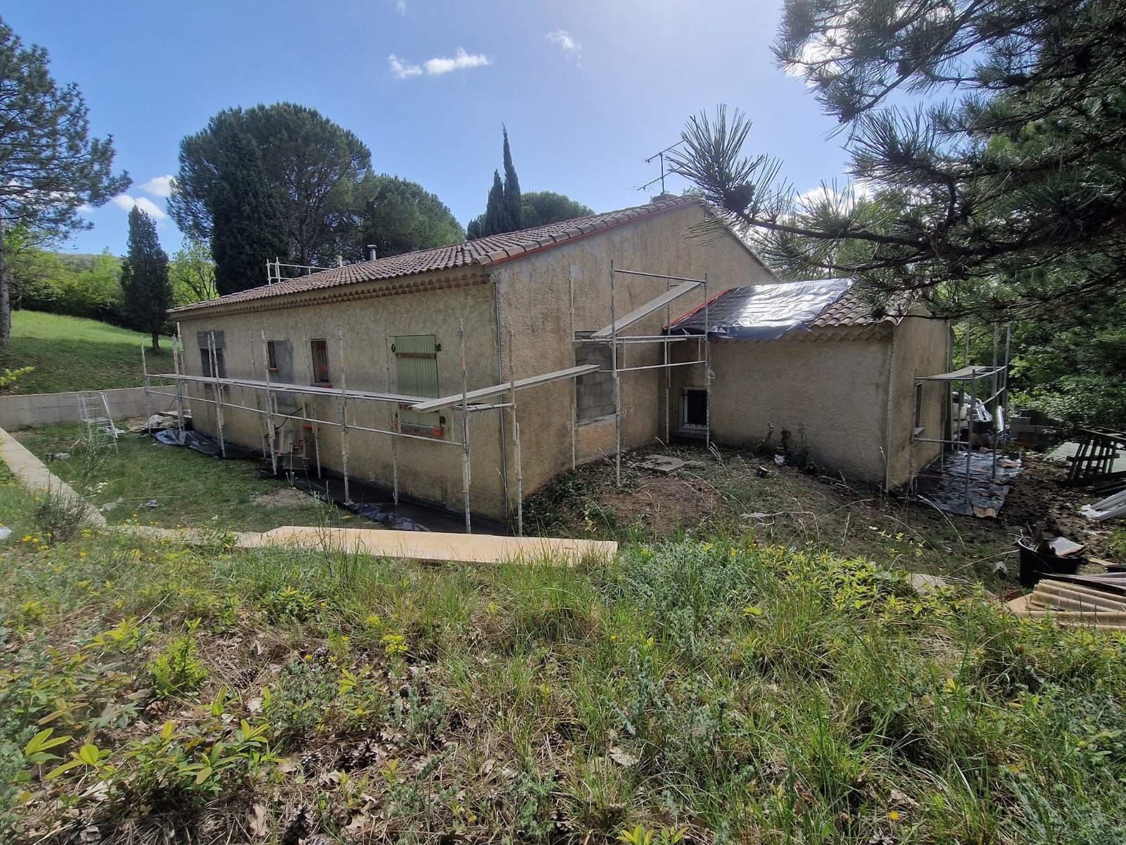 Maison beige en construction dans une cour herbeuse, entourée d'arbres.