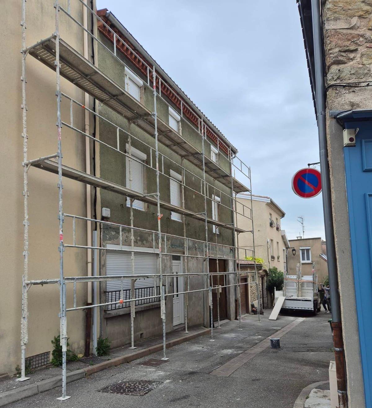 Un couvreur sur un échafaudage répare un toit bleu foncé. On aperçoit des bâtiments aux façades et fenêtres rouges et blanches.